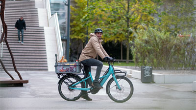 Zu sehen ist ein Mann auf einem blauen Cube Longtail Hybride Lastenrad, der durch die Innenstadt fährt und Lebensmittel gelagert hat.
