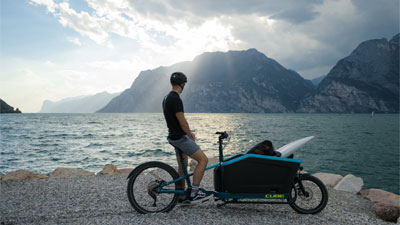 Ein Mann steht mit seinem Lastenrad, am Seeufer. Er betrachtet die Berge im Hintergrund.