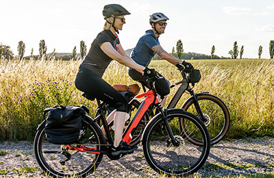 Zu sehen ist ein Paar welches mit E-Bikes über einen Schotterweg fährt. Im Hintergrund ist hohes Gras.