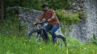Ein Radfahrer fährt mit seinem Mountainbike durch eine Wiese, im Hintergrund ist eine Mauer aus Stein.