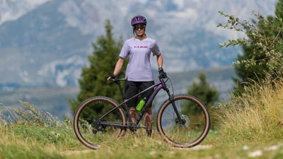 Zu sehen ist eine Frau in lila Fahrradbekleidung, vor ihr steht ein lila Cube Mountainbike. Im Hintergrund ist bergige Landschaft zusehen.