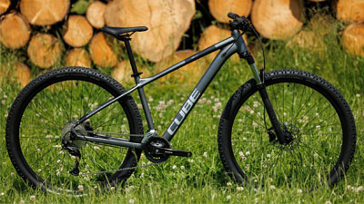 Zu sehen ist ein graues Cube Mountainbike, es steht auf einer Wiese. Im Hintergrund ist Holz gestapelt.