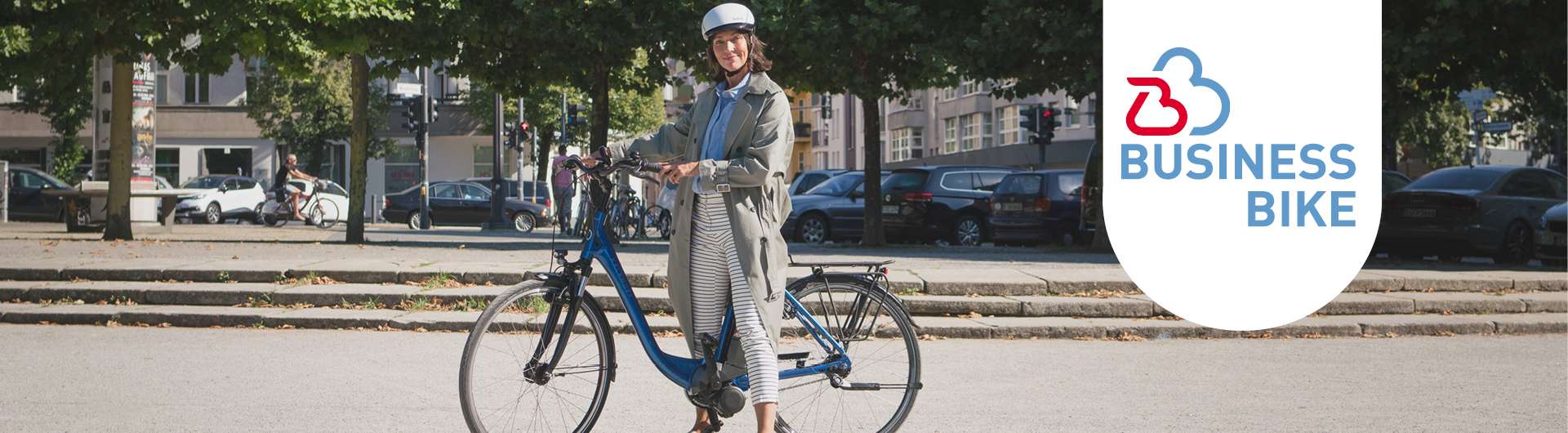 In einem Stadtpark steht eine Frau mit ihrem blauen Fahhrad. Neben ihr ist das Businessbike-Logo zu sehen.