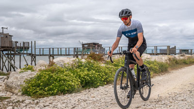Zu sehen ist ein Mann in Fahrradbekleidung. Er fährt sein Gravelbike auf einem Strandweg.