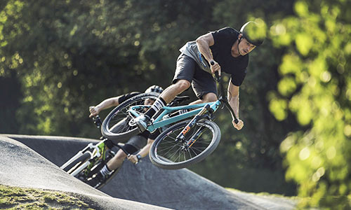 Radfahrer springt mit einem hellblauen Dirt Bike in einem Bikepark, ein weiterer Radfahrer mit grünem Fahrrad im Hintergrund.