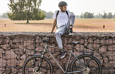 Zu sehen ist ein Mann mit Helm, welcher auf einer Mauer sitzt. Im Vordergrund steht ein graues Fahrrad und im Hintergrund ist eine Feldlandschaft.