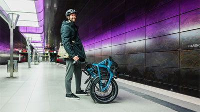 Zu sehen ist ein Mann in einer U-Bahn Station, vor ihm steht ein eingeklapptes blaues Faltrad.