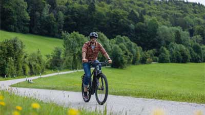 Ein Radfahrer fährt mit seinem Mountainbike über einen geteereten Radweg, im Hintergrund ist eine ländliche Landschaft.