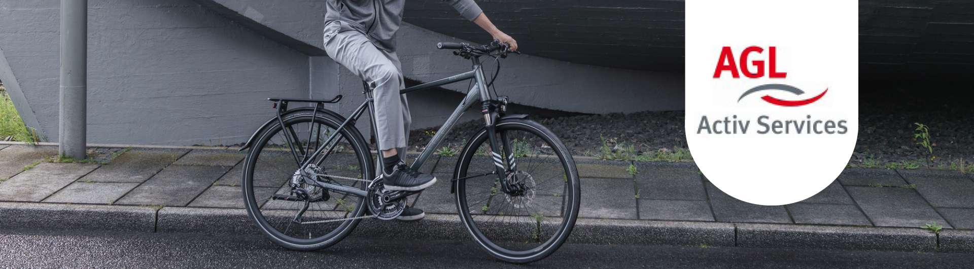 Ein Mann sitzt auf einem Fahrrad neben einem Gehweg. Neben ihm ist das AGL-Logo zu sehen.