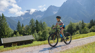 Ein Junge fährt auf seinem Mountainbike, auf einem Schotterweg, durch eine bergige Landschaft. Im Hintergrund ist eine Hütte zusehen.