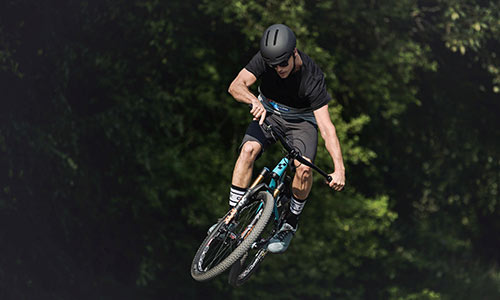 Radfahrer springt mit einem hellblauen Dirtbike, im Hintergrund sind Bäume zu sehen.