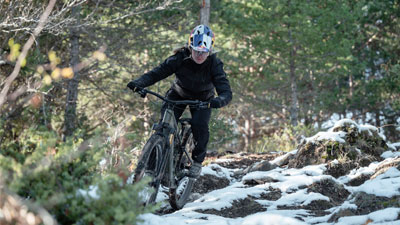 Eine Radfahrerin fährt mit ihrem Mountainbike, durch den unebenen Wald, teilweise ist der Boden noch mit Schnee bedeckt.